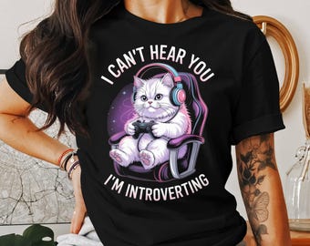 Puede incluir: Camiseta negra con un gato blanco de dibujos animados con auriculares y jugando a un videojuego. El gato está sentado en una silla de juego. El texto de la camiseta dice "I CAN'T HEAR YOU I'M INTROVERTING."
