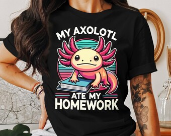 Puede incluir: Camiseta negra con un ajolote rosa y amarillo sosteniendo un libro. El texto en la camiseta dice "Mi ajolote se comió mi tarea."