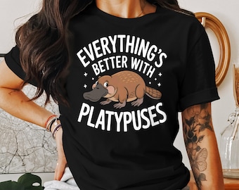May include: Black t-shirt with the text "EVERYTHING'S BETTER WITH PLATYPUSES" in white. The shirt features a cartoon platypus in shades of brown and gray. The design includes small star accents.