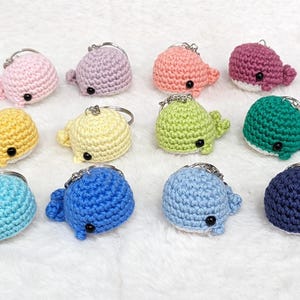 May include: A collection of 12 crocheted whale keyrings in various colours, including pink, purple, orange, green, yellow, blue, and light blue.