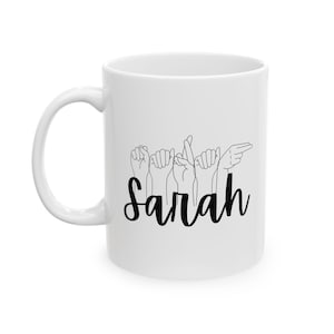 Peut inclure: Une tasse en céramique blanche avec un motif noir de mains signant le nom "Sarah".