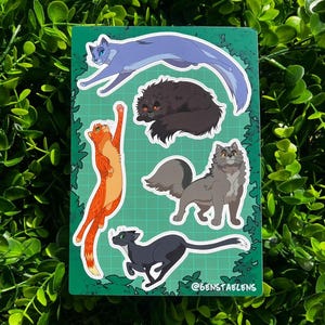 May include: A sticker sheet featuring five cartoon cat illustrations in various poses and colors. The cats include a blue cat leaping, a black cat curled up, an orange cat reaching up, a gray cat standing, and a black cat running. The background is green with a grid pattern.