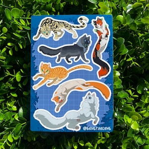May include: A sticker sheet featuring seven stylized cat illustrations in various poses. The cats are depicted in shades of gray, orange, brown, and white, with blue and green eyes. The background is a blue grid pattern. The text "@6ENSTAELENS" is at the bottom.