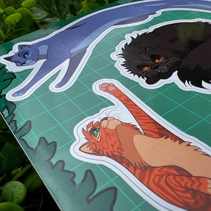 May include: A sheet of stickers featuring three stylized cat illustrations. One is a long, blue cat with blue eyes. Another is a black cat with orange eyes. The third is an orange tabby cat with green eyes, stretching upwards. The background is a green grid.