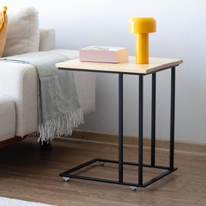 May include: A modern side table with a light wood top and black metal frame. A yellow lamp and a pink box sit on the table. The table is next to a white sofa with a gray blanket.