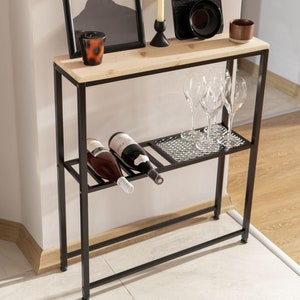 May include: Black metal console table with a wood top shelf, two lower shelves, and a wine rack. The table has a framed picture, a candle, a tray, and wine glasses on top.