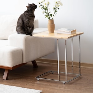 May include: A modern side table with a light wood top and chrome legs. A vase of white flowers sits on the table, next to a book. A dark gray cat sits on a white sofa, looking at the flowers.