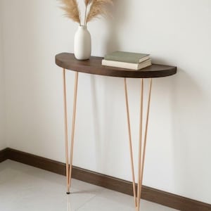 May include: A half-moon console table with a dark wood top and copper-coloured hairpin legs. A white vase with dried pampas grass and two books are on the table. The table is approximately 76 cm long.