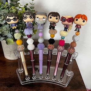 Puede incluir: Siete bolígrafos temáticos de Harry Potter con cuentas de colores y pedrería. Los bolígrafos se muestran en un soporte de acrílico transparente.