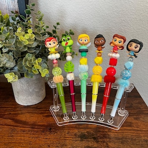 May include: Set of six colourful pens with different cartoon character toppers. Each pen has a gradient colour and is decorated with beads and charms. The pens are displayed in a clear plastic stand.
