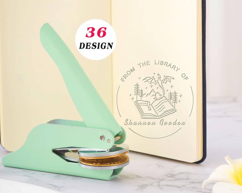 May include: A mint green handheld embosser tool positioned next to an open notebook. The notebook page displays a circular stamp design featuring a dragon, mountains, trees, and the text "FROM THE LIBRARY OF Shannon Gooden". A "36 DESIGN" label is visible.