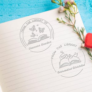 May include: Two circular embossed stamps on a lined notebook page. The first stamp features an open book with three butterflies and the text FROM THE LIBRARY OF Shannon Gooden. The second stamp shows an open book with a cup and glasses.