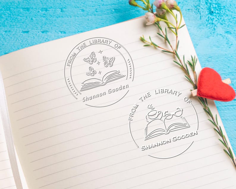 May include: Two circular embossed stamps on a lined notebook page. The first stamp features an open book with three butterflies and the text FROM THE LIBRARY OF Shannon Gooden. The second stamp shows an open book with a cup and glasses.