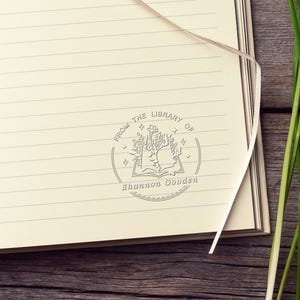 May include: A cream-colored lined notebook page featuring a circular stamp with an open book illustration, floral elements, and stars. The text reads FROM THE LIBRARY OF and Shannon Gooden. A thin ribbon bookmark rests on the page.
