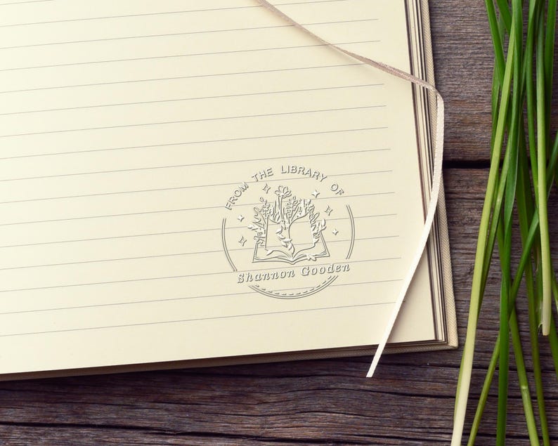 May include: A cream-colored lined notebook page featuring a circular stamp with an open book illustration, floral elements, and stars. The text reads FROM THE LIBRARY OF and Shannon Gooden. A thin ribbon bookmark rests on the page.