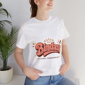 May include: A white short-sleeve t-shirt featuring a graphic design with the word Bindass in a bold, orange-red retro font. Above the text is a small orange crown, and below it, the phrase carefree / unapologetically bold is printed in black.