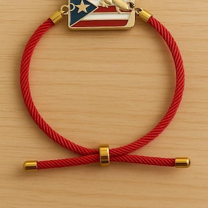 May include: Red rope bracelet with gold-colored accents. The centerpiece features a rectangular charm with the Puerto Rican flag and a gold-colored rocket. The bracelet has a sliding knot closure.