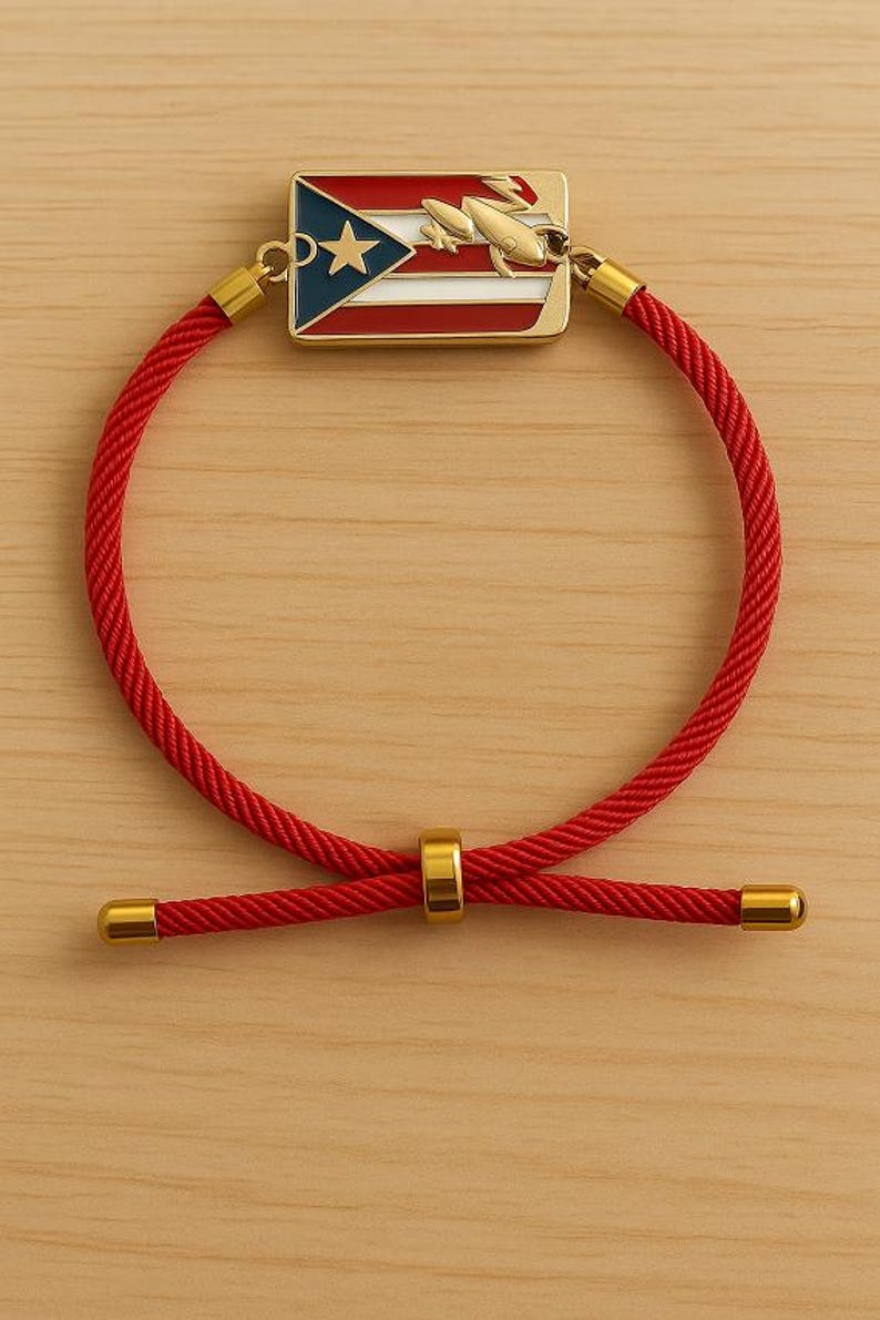 May include: Red rope bracelet with gold-colored accents. The centerpiece features a rectangular charm with the Puerto Rican flag and a gold-colored rocket. The bracelet has a sliding knot closure.
