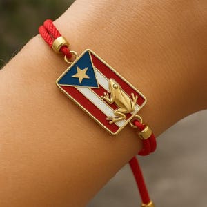 May include: Red cord bracelet featuring a gold-colored charm with the Puerto Rican flag and a gold frog. The charm has a blue triangle with a star, white stripes, and red stripes. The bracelet has gold-colored accents.