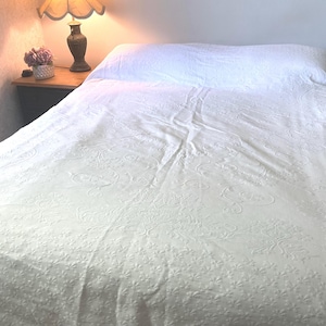 May include: A white bedspread featuring an intricate, tone-on-tone embossed floral and scrollwork pattern covering the entire surface. The fabric has a textured appearance with a repeating botanical design that creates a subtle, raised effect.