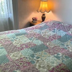 May include: A quilted bedspread featuring a patchwork pattern of floral and geometric designs in shades of magenta, teal, sage green, and cream. The fabric has a diamond-stitched texture throughout, covering the surface of a bed.