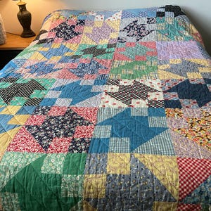 May include: A patchwork quilt featuring a repeating star pattern composed of various fabric squares in diverse colours and prints, including red, blue, green, yellow, and black, with visible stitching throughout the entire surface of the textile.