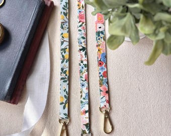 May include: Three floral patterned lanyards with gold and silver swivel clasps. The lanyards feature various floral designs with yellow, pink, and blue flowers on a light background. The lanyards are displayed on a cream-colored surface.