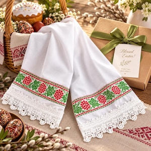 May include: A white linen towel featuring a decorative border with red and green floral embroidery and a scalloped white lace trim. The fabric is folded, showcasing the intricate needlework and the delicate lace edging along the bottom hem.