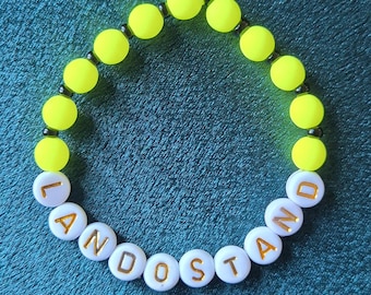 Puede incluir: Una pulsera de cuentas con la palabra "LANDOSTAND" escrita con cuentas de letras blancas con letras doradas. La pulsera también presenta cuentas de color amarillo brillante. La pulsera está sobre un fondo turquesa.