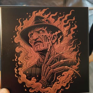 May include: A black and copper coloured ceramic tile featuring a graphic of Freddy Krueger from the Nightmare on Elm Street film series. Freddy is shown wearing his trademark fedora, clawed glove, and a menacing expression. He is surrounded by flames.