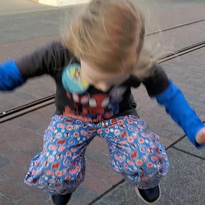 May include: A child jumping, wearing a black long-sleeved top with a Spider-Man graphic, blue and red patterned trousers, and black shoes. The child has blonde hair and is in motion.