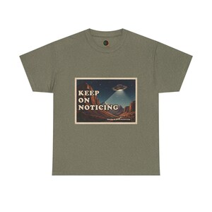 May include: An olive green t-shirt featuring a rectangular graphic print of a desert landscape at night with a flying saucer emitting a beam of light. The text "KEEP ON NOTICING" is printed in white letters on the left side of the graphic.