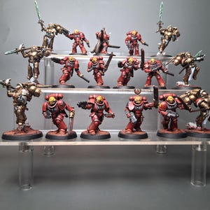 Puede incluir: Figuras en miniatura de soldados con armadura roja y dorada, posando con armas. Las figuras se exhiben en estantes de acrílico transparente. Algunas figuras sostienen espadas, otras armas de fuego.
