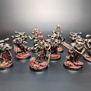 May include: A group of miniature figurines of armoured warriors. The figures are painted in metallic colours with red and green accents, standing on textured, reddish-brown bases. The theme suggests a fantasy or sci-fi tabletop game.