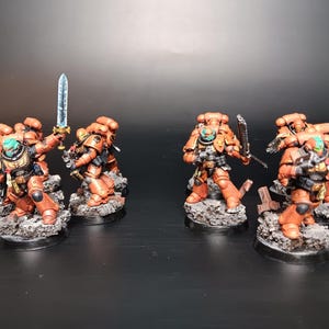 May include: Miniature figurines of armored soldiers in orange and teal, posed on rocky bases. One figure holds a glowing blue sword aloft. The figures are detailed with weapons and gear, set against a dark background.