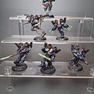 Op de afbeelding: Miniatuurfiguren van gepantserde figuren in blauw en goud, met groene klauwachtige handen en wapens. De figuren staan op een gelaagde display, die hun gedetailleerde ontwerpen en dynamische houdingen laat zien.