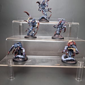 Puede incluir: Cinco figuras en miniatura de una criatura con cuerpo segmentado, garras afiladas y cola larga. Las figuras están pintadas en tonos de azul, gris y rojo, y se exhiben en estantes de acrílico transparente. Las figuras están posadas en varias posturas de acción.