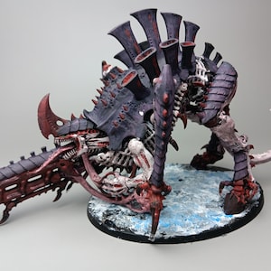 Puede incluir: Una detallada miniatura de una criatura Tiránida, con una coraza gris oscuro y detalles en rojo y color hueso. El modelo está posado sobre una base texturizada, mostrando detalles intrincados y un diseño amenazante.