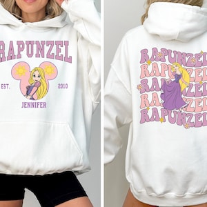 May include: White hoodie with a purple Rapunzel graphic on the front and a repeated Rapunzel graphic on the back. The front graphic features Rapunzel in a Mickey Mouse ear silhouette with the text "EST. 2010" and "JENNIFER".