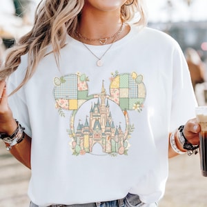 Könnte beinhalten: Weißes T-Shirt mit einem Mickey Mouse Kopf-Design, mit einem Schloss in der Mitte. Das Design verwendet Pastellfarben, florale Details und Ostereier. Ein lässiges Kleidungsstück.
