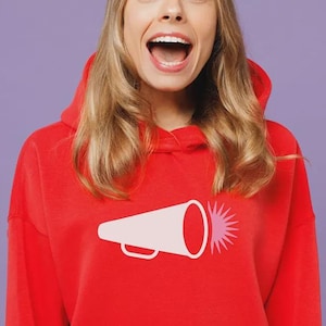 May include: A bright red long-sleeve hooded sweatshirt featuring a graphic print on the chest. The graphic depicts a light pink megaphone with a dark pink starburst pattern emanating from the front opening of the megaphone speaker.