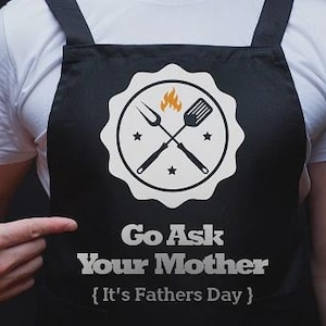 May include: A black apron featuring a white circular emblem with a flame, a fork, and a spatula. Below the emblem, the text reads: Go Ask Your Mother { It's Fathers Day }. The apron has black straps and is worn over a plain white t-shirt.