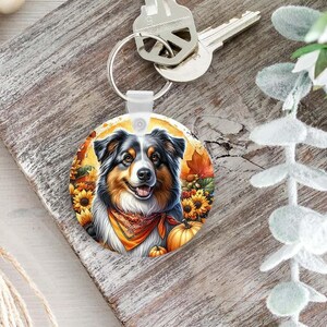 May include: A round keychain featuring a colorful illustration of an Australian Shepherd dog wearing an orange bandana, surrounded by autumn sunflowers, fall leaves, and pumpkins, attached to a silver metal key ring with a metal key.