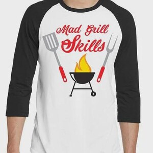 May include: A white baseball t-shirt with black three-quarter length sleeves featuring a graphic of a grill with a flame, a spatula, and a fork. The text "Mad Grill Skills" is printed in red above the grill graphic in a stylized font.