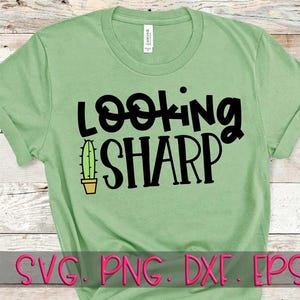 May include: A light green short-sleeved t-shirt featuring the black text "LOOKING SHARP" with a small green cactus in a tan pot replacing the letter "I". The shirt has a crew neckline and a standard fit, displaying a graphic design on the front.