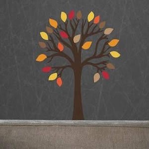 May include: A wall decal featuring a brown tree trunk and branches with multi-coloured leaves in shades of yellow, orange, red, and tan. The tree design is applied to a dark grey textured wall surface positioned above a light grey upholstered sofa.