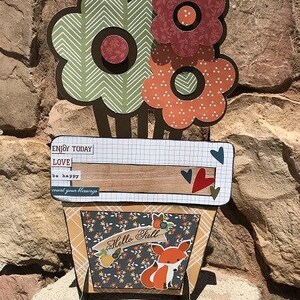 May include: A decorative paper craft display featuring three layered flowers in green and orange patterns atop a rectangular base. The base includes the text ENJOY TODAY, LOVE, be happy, count your blessings, and Hello Fall with a fox illustration.