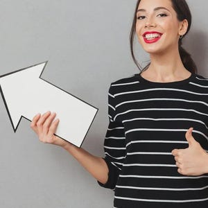May include: A black long-sleeved shirt with horizontal white stripes, featuring a solid white arrow-shaped sign with a black border held in one hand, while the other hand displays a thumbs-up gesture against a neutral gray background.