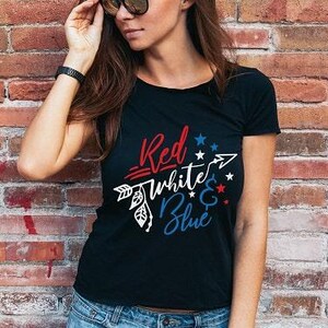 May include: A black short-sleeved t-shirt featuring a graphic design with an arrow and the text "Red White & Blue" in red, white, and blue colours. The design includes small star accents and decorative leaf patterns integrated into the arrow shaft.