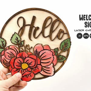 Puede incluir: Un letrero de bienvenida de madera con la palabra "Hello" en fuente cursiva. El letrero es redondo con un borde marrón oscuro y presenta flores rojas y rosas con hojas verdes. También se ve el texto "WELCOME SIGN".